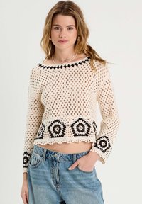 Top de ganchillo en color crema con acentos florales en negro y dobladillo con forma de concha; cuenta con mangas largas y un diseño abierto y aireado, combinado con pantalones de mezclilla.