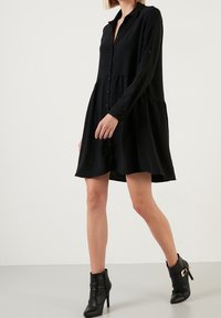 Abito camicia nero a maniche lunghe con bottoni davanti, design a livelli e tessuto morbido. Indossato con stivali neri alla caviglia con una fibbia.
