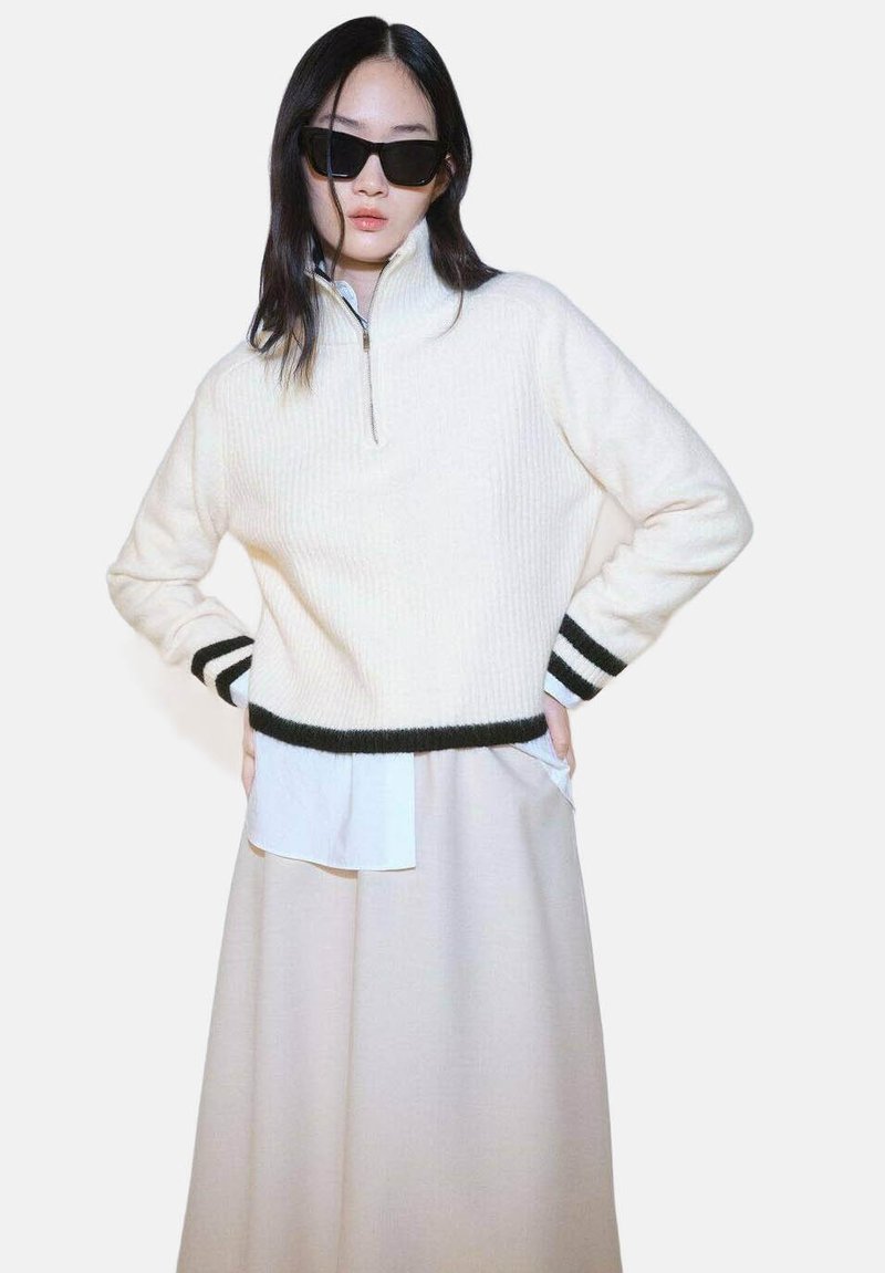 Cream-colored sweater with a high collar, ribbed texture, and black stripe accents at sleeves. Worn over a white shirt and beige skirt.