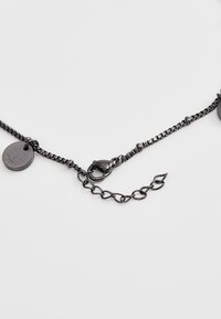 Schwarze Metallkette mit einer langen Kette im strukturierten Gliederdesign, einem Verschluss und einem runden Anhänger mit geprägtem Logo.