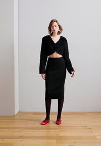 Giacca nera corta e pelosa con maniche lunghe, abbinata a una gonna a matita nera a coste e scarpe rosse lucide. Sfondo semplice e pavimento in legno.
