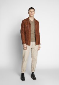 Chaqueta de cuero marrón con cierre de botones en el frente y dos bolsillos en el pecho, combinada con un suéter beige, pantalones claros y botas negras de tobillo.