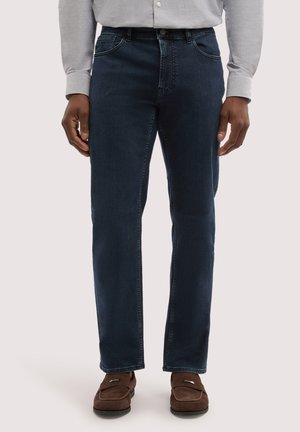 Uomo che indossa jeans blu scuro, maglia a maniche lunghe grigio chiaro e mocassini in pelle scamosciata marrone, in piedi davanti a uno sfondo uniforme.