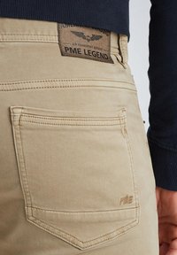 Pantalones de algodón beige con un corte ajustado, que cuentan con una etiqueta distintiva, bolsillos traseros con costuras en contraste y detalle de logo en relieve.
