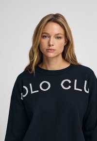 Svart tröja med lös passform, ribbat halslinne och vit "POLO CLUB" text över bröstet. Mjuk textur och sportig design.