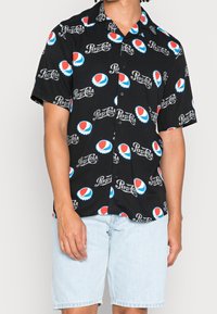 Short-sleeve black button-up shirt featuring a repeating Pepsi logo pattern in red, white, and blue on a smooth fabric. Paired with denim shorts.