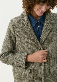 Manteau en tweed texturé vert et crème avec un col, des manches longues, deux poches avant et fermeture à bouton. Porté sur une chemise en denim.