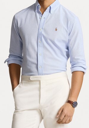 Mann trägt hellblau gestreiftes Hemd mit hochgekrempelten Ärmeln, weiße Hose und braune Lederarmbanduhr am linken Handgelenk.