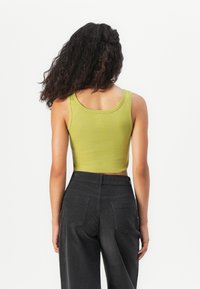 Woman with long curly hair wears a green sleeveless crop top and high-waisted black pants, standing with her back to the camera.