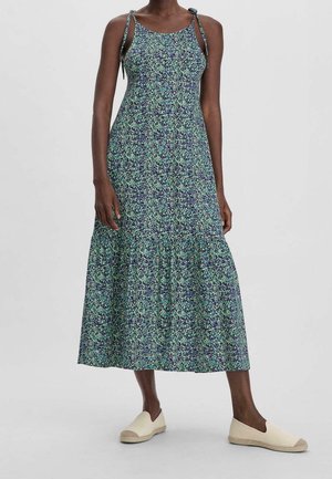 Kvinde iført en ærmeløs blomsterkjole, der går til anklerne, med bindestropper og hvide slip-on sko, stående mod en ensfarvet baggrund.
