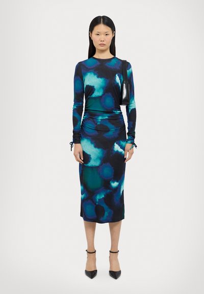 Robe à manches longues dans un motif tie-dye bleu-vert, présentant une silhouette ajustée, des détails froncés, et accompagnée de talons noirs à bride de cheville.