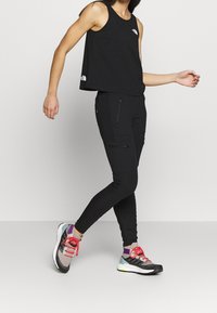 Femme marchant dans un haut sans manches noir et un pantalon cargo noir, portant des chaussures de course multicolores avec des lacets roses sur un fond uni.