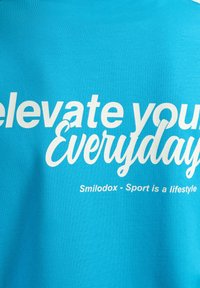 Turquoise T-shirt met grote witte tekst: "Verhoog je dagelijkse leven" en "Smilodox - Sport is een levensstijl." Zachte stof, relaxte pasvorm.