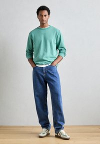 Mäta zelená mikina, modré pohodlné džínsy a tenisky s green pruhmi. Neformálny outfit; textilná látka, jednoduchý dizajn a tlmené farby.