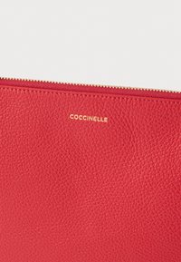 Piros texturált bőr erszény arany színű cipzárral és a "Coccinelle" dombornyomott logóval az elején.