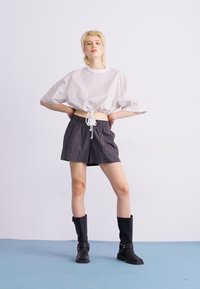 Witte en grijs gestreepte oversized top gecombineerd met zwart gestreepte shorts. Model draagt hoge zwarte laarzen met gespen en staat op een blauwe ondergrond.