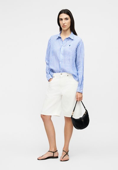 Femme portant une chemise à manches longues rayée bleu et blanc, un short blanc jusqu'au genou, des sandales noires et tenant un sac à main noir.