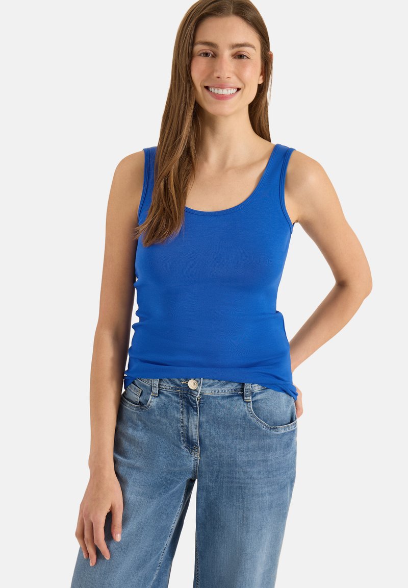 Femme souriante portant un débardeur bleu sans manches et un jean bleu clair, debout avec une main dans le dos.