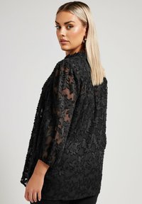 Blouse noire en voile avec broderie florale, manches trois-quarts et coupe décontractée. Le design texturé ajoute de la profondeur et des détails.