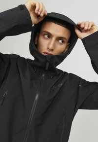 Schwarze wasserdichte Jacke mit Kapuze, ausgestattet mit einem Frontreißverschluss, Seitentaschen und einem eleganten Design. Das Material wirkt glatt mit einer matten Oberfläche.