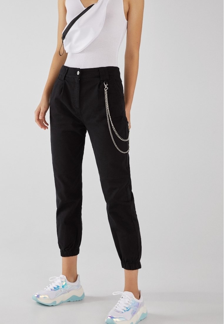 Pantalons noirs fuselés avec des poignets élastiques, dotés de poches latérales et d'un accent en chaîne argentée. Portés avec un débardeur blanc et des baskets colorées.