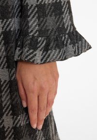 Gray and black houndstooth-patterned fabric with a ruffled cuff. The hand rests on the edge, showcasing the texture and pattern details.