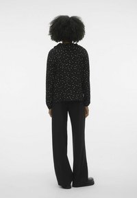 Frau, die mit dem Rücken zum Betrachter steht, trägt weite schwarze Hosen und ein schwarzes Langarmshirt mit Punkten sowie Locken.