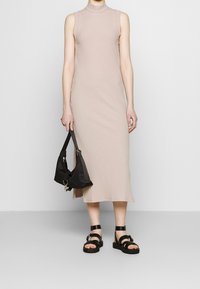 Abito midi senza maniche a coste beige con colletto alto, abbinato a una borsa a spalla nera e sandali neri con cinturini. Tessuto liscio, design aderente.