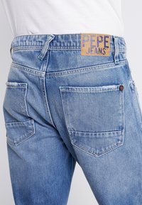 Vaaleansiniset farkut, joissa on kaksi takataskua ja nahkalaatta, jossa lukee "PEPE JEANS", yhdistettynä valkoiseen paitaan.