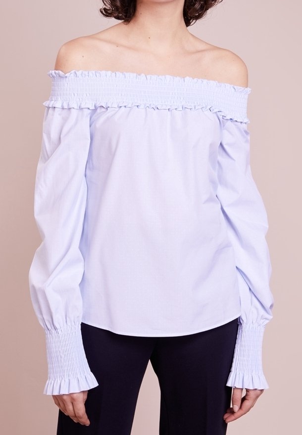Femme portant un chemisier bleu clair à épaules dénudées avec de longues manches bouffantes et des poignets froncés, associé à un pantalon foncé.