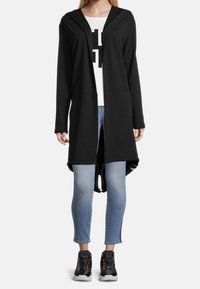 Chaqueta larga con capucha negra hecha de una tela suave, con abertura frontal, bolsillos laterales y un dobladillo curvado. Se lleva con jeans azul claro y zapatillas negras.