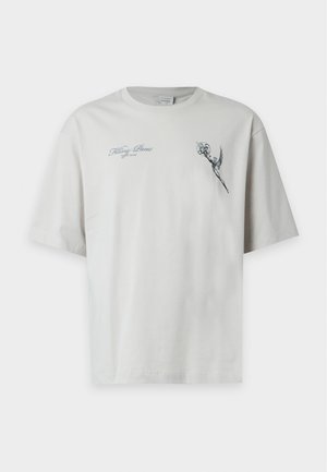 Jeune homme debout avec une main dans la poche, portant un T-shirt gris clair avec un motif floral et un pantalon large noir ainsi que des chaussures.
