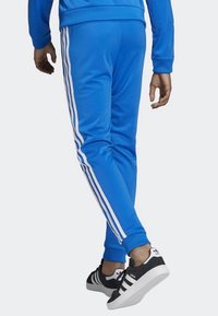 Modré joggingové kalhoty vyrobené z hladkého materiálu, s třemi bílými pruhy po stranách, s užším střihem a žebrovanými manžetami. Jsou obuty černé tenisky.