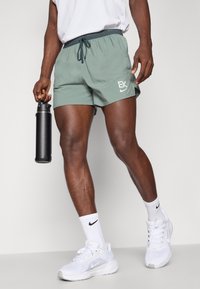 Pantalones cortos deportivos verdes con cintura elástica y cordón, con un logo blanco. La persona sostiene una botella de agua negra y lleva zapatillas blancas.