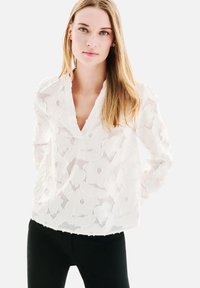 Jeune femme aux longs cheveux blonds portant un haut en dentelle florale blanche et un pantalon noir, devant un fond uni.