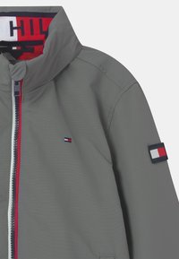Šedá voděodolná bunda s vysokým límcem, předním zipem s červenými akcenty a logem na rukávu s červenými, bílými a modrými detaily.