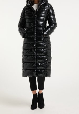 Long manteau matelassé noir brillant avec capuche, porté avec un pantalon noir et des bottines noires, mains dans les poches, debout sur un fond blanc.