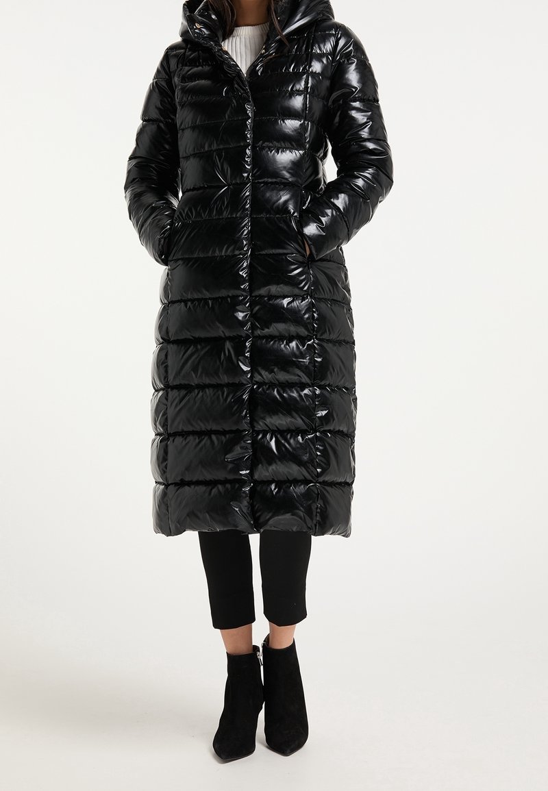 Long manteau matelassé noir brillant avec capuche, porté avec un pantalon noir et des bottines noires, mains dans les poches, debout sur un fond blanc.