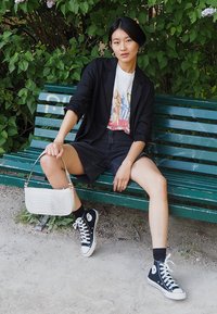 Schwarze, maßgeschneiderte Blazer über einem Grafik-T-Shirt, Jeans-Shorts, weiße Handtasche mit Krokodil-Prägung, schwarze High-Top-Sneaker mit weißen Sohlen.