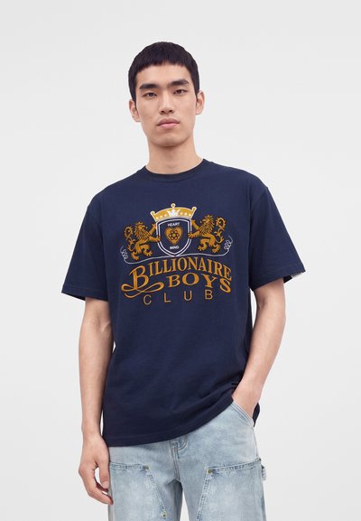 Jeune homme portant un t-shirt bleu marine Billionaire Boys Club avec un écusson doré et un jean bleu clair, debout devant un fond uni.