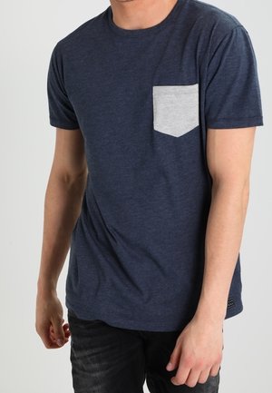 Homme portant un t-shirt bleu marine à manches courtes avec une poche poitrine gris clair et un pantalon noir, debout devant un fond uni.