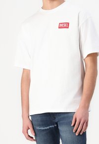 Vit bomull kortärmad T-shirt med en röd rektangulär "DIESEL"-logotyp på vänster bröst. Kombineras med blå slitna denimjeans.