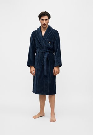 Homme debout pieds nus portant un peignoir en peluche bleu foncé noué à la taille avec des poches avant et un petit ours brodé sur la poche poitrine.