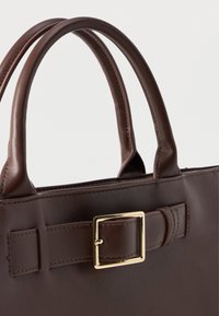 Bolsa de pele castanha com duas alças arredondadas, um detalhe em fivela quadrada dourada e costura decorativa no corpo.