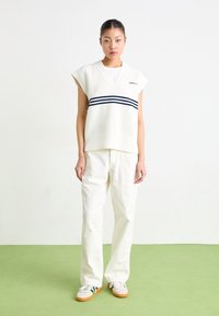 White sleeveless sweater with a V-neck and navy stripes, paired with loose white pants and casual sneakers. Smooth texture, modern design.