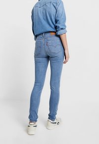 Jeans de mezclilla de color azul claro con un corte ajustado, que presentan bolsillos traseros y un parche de cuero marrón en la cintura. Combinados con zapatillas blancas.