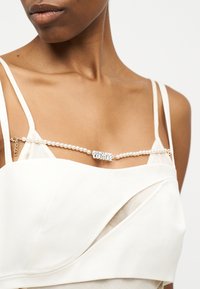 Γυναίκα που φοράει λευκό τοπ με τιράντες και μαργαριταρένια αλυσίδα με ασημένιο λογότυπο "Versace Jeans Couture" στο στήθος.