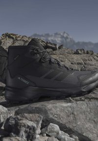 Black Adidas Terrex hiking boot resting on rocky terrain with blurred mountain range in background under clear sky.