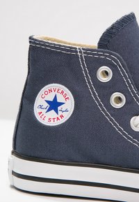 Dunkelblaue Canvas-Hochtop-Sneaker mit weißer Gummisohle, ausgestattet mit einem runden Logopatch mit einem Stern und Text, silbernen Ösen und kontrastierenden Nähten.