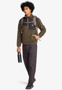 Brown fleece jacket with black chest pocket and matching straps; dark trousers and black sneakers; holding a black water bottle.